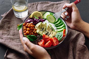 Eine Bowl mit Avocado, Rohkost und Dip. Eine Frauenhand hält eine Gabel, auf die sie eine Gurkenscheibe gespießt hat.