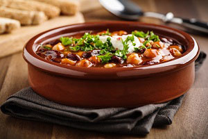 Auf einem Holztische steht herzhaftes Chili, in einer braunen Keramikschale angerichtet und mit Petersilie garniert. Im Hintergrund liegt ein Löffel und frisch aufgeschnittenes Brot.