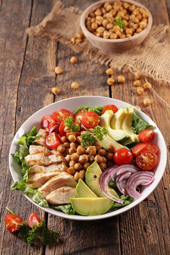 Eine Bowl mit Hähnchenfleisch, Kichererbsen und Rohkost, appetitlich auf einem Holztisch angerichtet